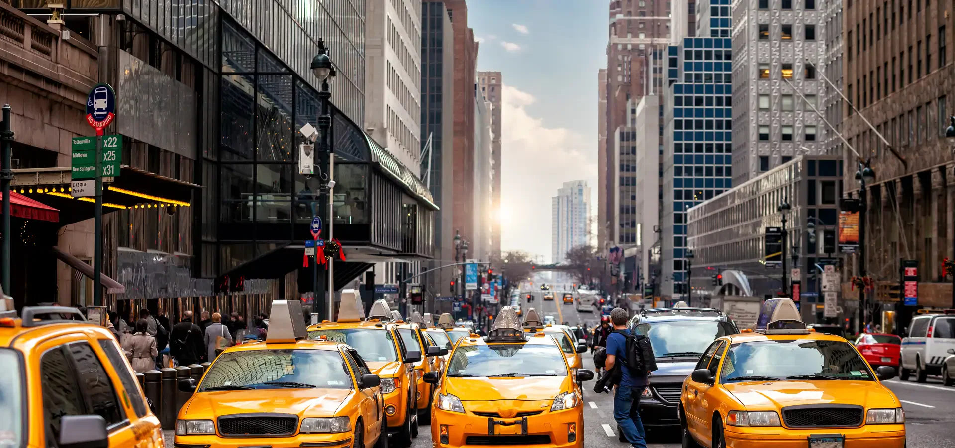 Busy city street filled with yellow taxis, pedestrians, and modern buildings under a clear sky, with the sun setting in the distance.