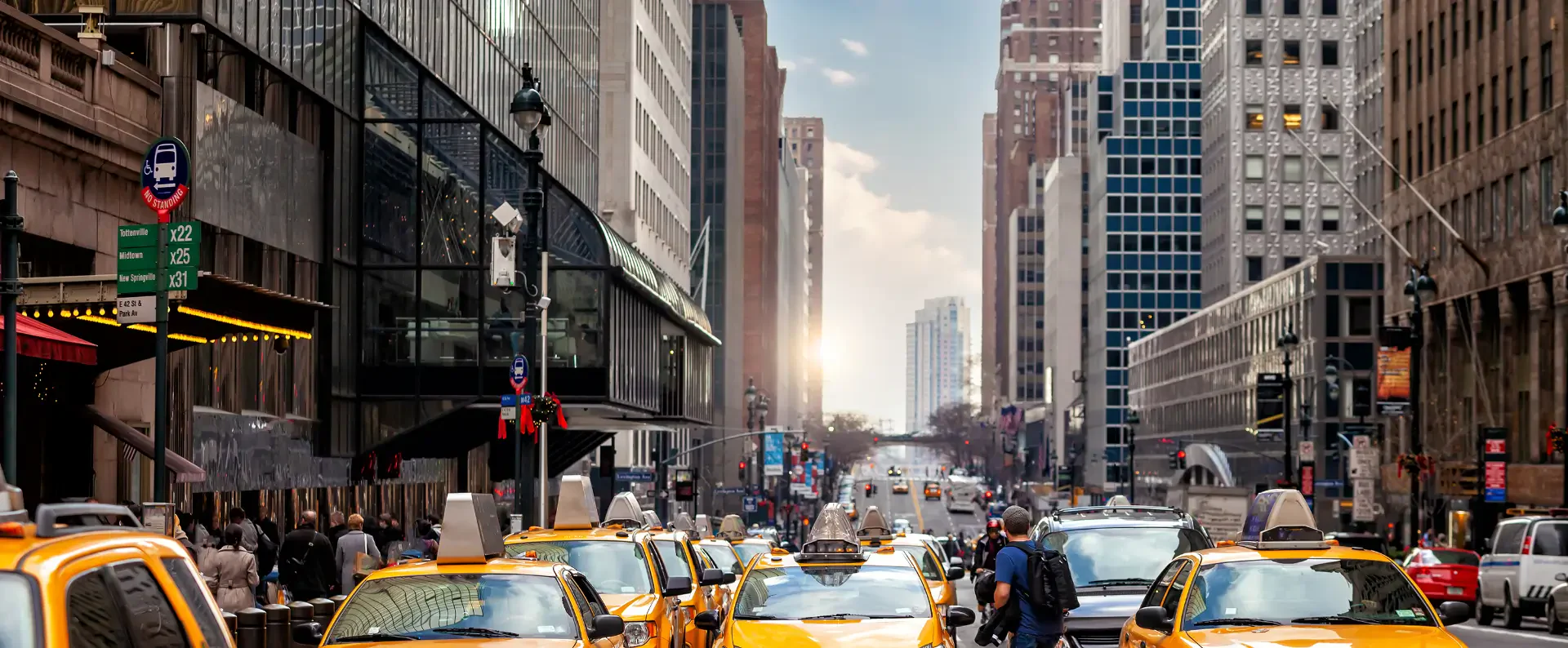 Busy New York City street lined with yellow taxis, bustling pedestrians, glass buildings, and a glowing sunset in the background.